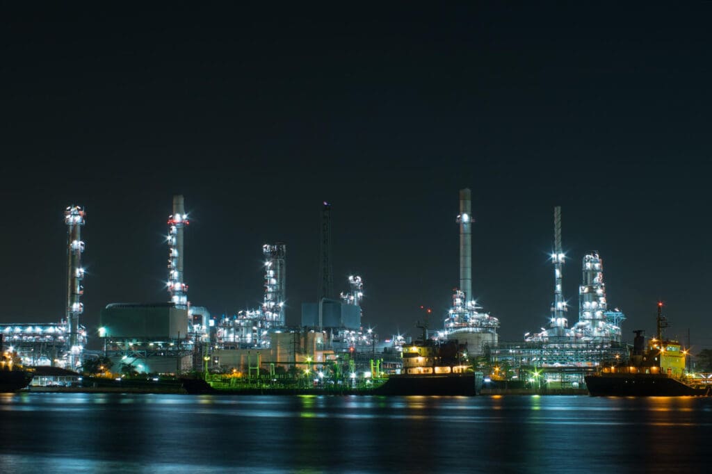 Refinaria industrial iluminada à noite, com torres, chaminés e navios atracados, representando operações integradas e alta capacidade no setor de óleo e gás.