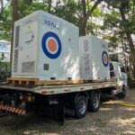 Sistema BESS da Ion Energia sendo transportado em caminhão, instalado em área arborizada com edifícios ao fundo.