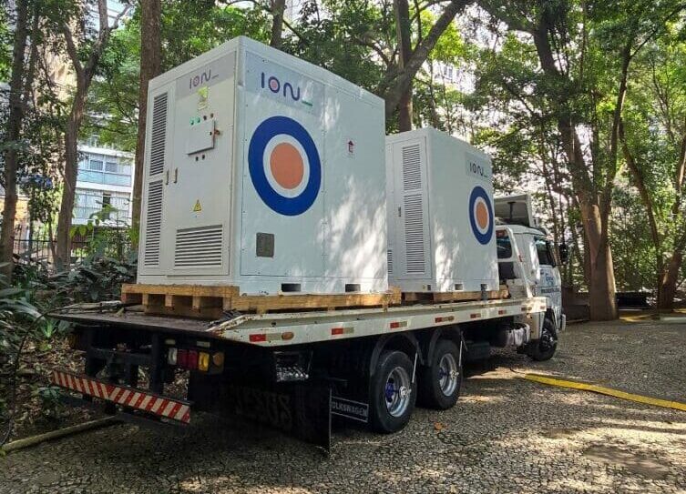 Sistema BESS da Ion Energia sendo transportado em caminhão, instalado em área arborizada com edifícios ao fundo.