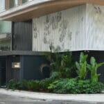 Fachada de edifício contemporâneo com revestimento metálico perfurado branco, destacando padrões geométricos e vegetação paisagística na entrada.