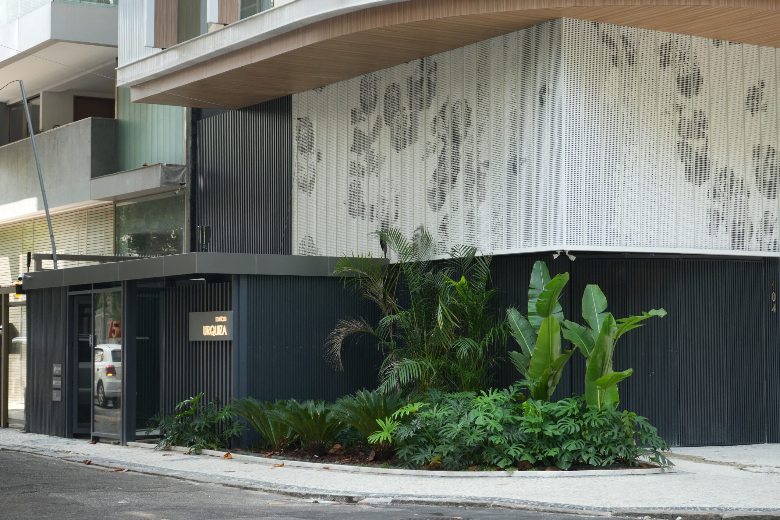 Fachada de edifício contemporâneo com revestimento metálico perfurado branco, destacando padrões geométricos e vegetação paisagística na entrada.