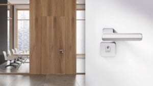 Maçaneta moderna da Linha Domus La Fonte em porta de escritório de madeira, com sala de reuniões ao fundo, representando design sofisticado e funcionalidade para ambientes profissionais.