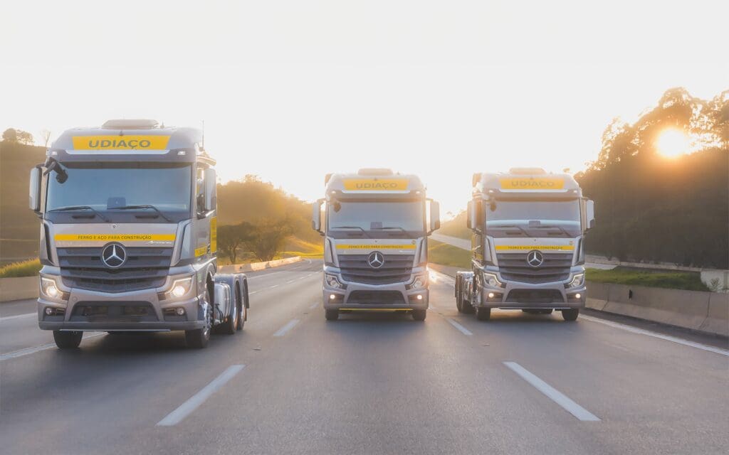Três caminhões da frota Udiaço em rodovia, prontos para entrega de aço para construção civil.