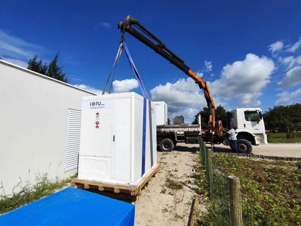 Sistema BESS da Ion Energia sendo instalado com guindaste em área externa, ao lado de um caminhão e sob céu azul.