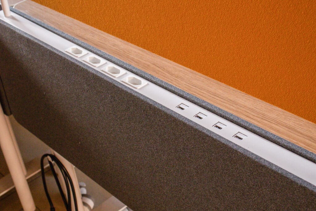 Detalhe de estação de trabalho Novara com tomadas elétricas e entradas USB embutidas em painel de madeira e tecido.