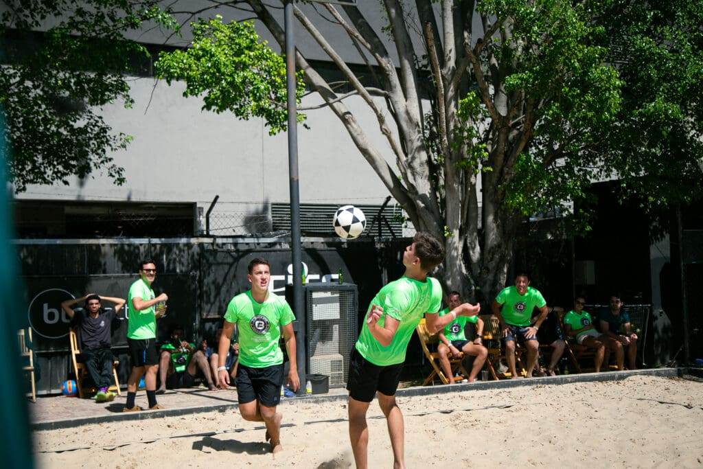 Atletas com camisetas verdes disputam partida de futevôlei em quadra de areia, enquanto outros participantes assistem ao fundo durante o 3º C3 Open Beach Sports.