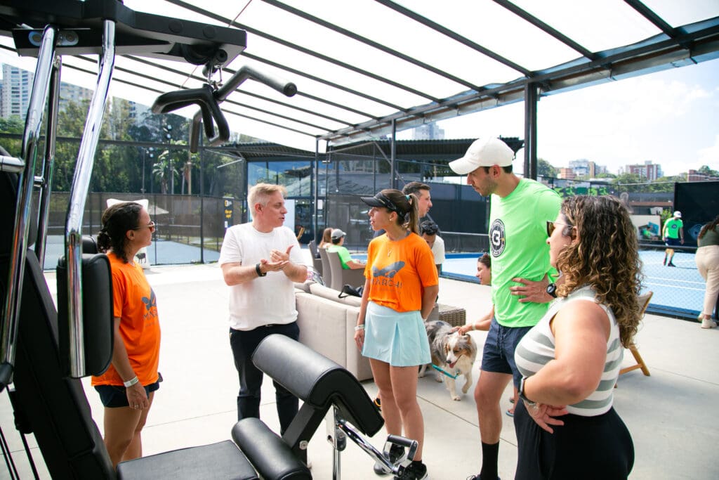 Grupo de participantes do 3º C3 Open Beach Sports conversa em área de convivência da Arena BTG, com quadras esportivas ao fundo e clima descontraído.
