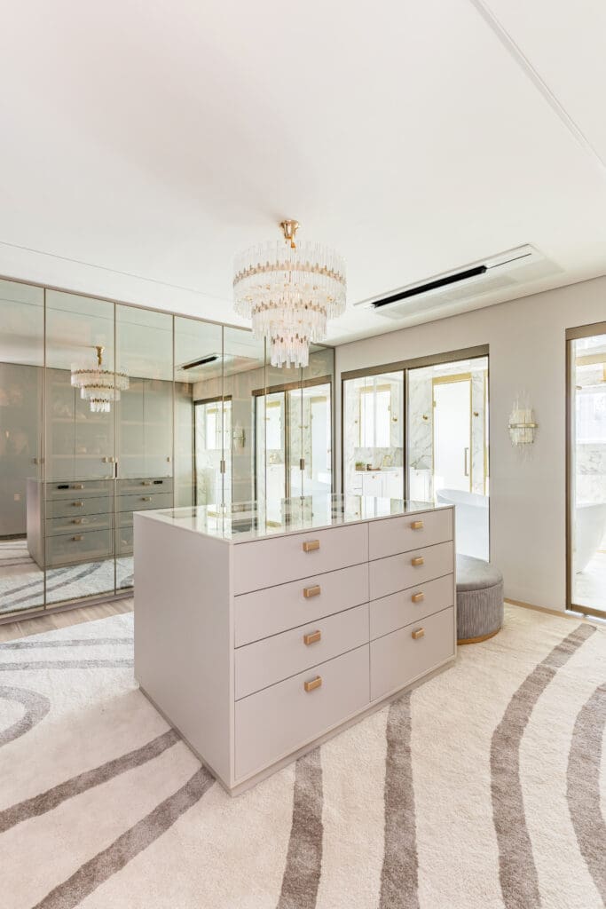 Ilha central com gavetas em walk-in closet sofisticado, armários espelhados e lustre de cristal, integrado ao banheiro.