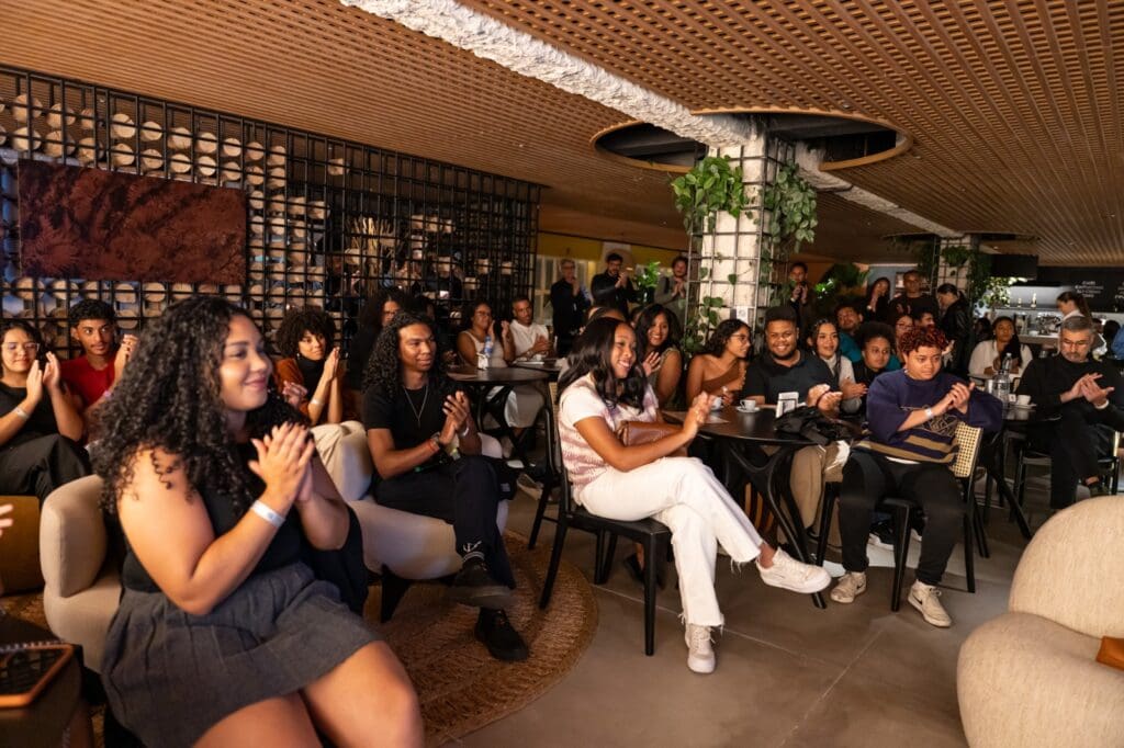 Público diverso de jovens e adultos aplaudindo durante evento do Programa Aproxima, promovendo inclusão étnico-racial na arquitetura.