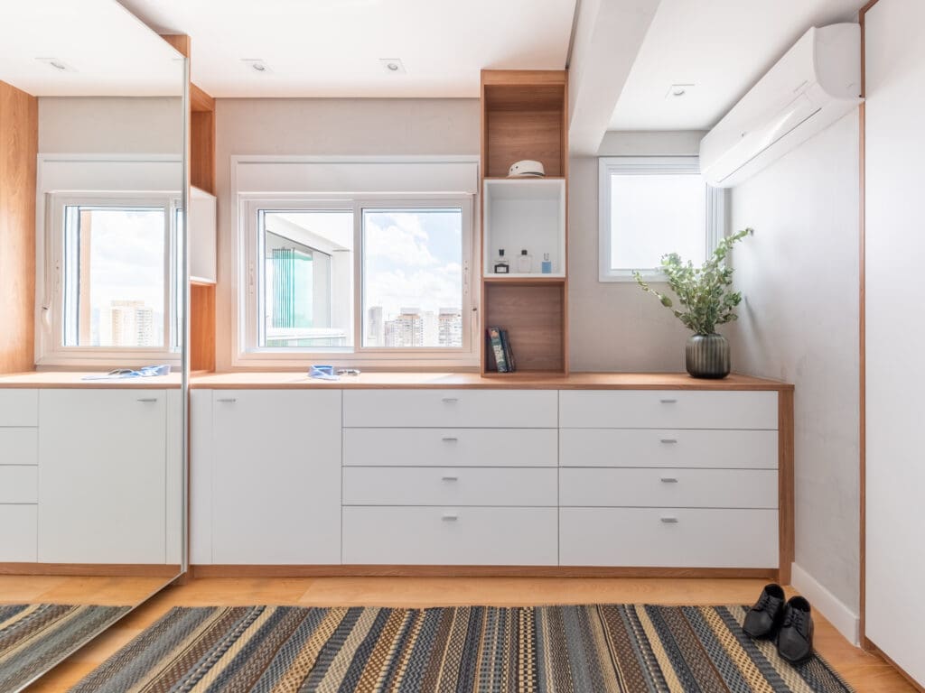 Walk-in closet moderno com marcenaria planejada em tons de branco e madeira, gaveteiros amplos, espelhos verticais, nichos decorativos, janelas com vista urbana, piso de madeira e tapete listrado