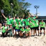 Grupo de atletas com camisetas verdes do 3º C3 Open Beach Sports posa sorridente e comemorativo na quadra de areia, celebrando o espírito esportivo do evento.