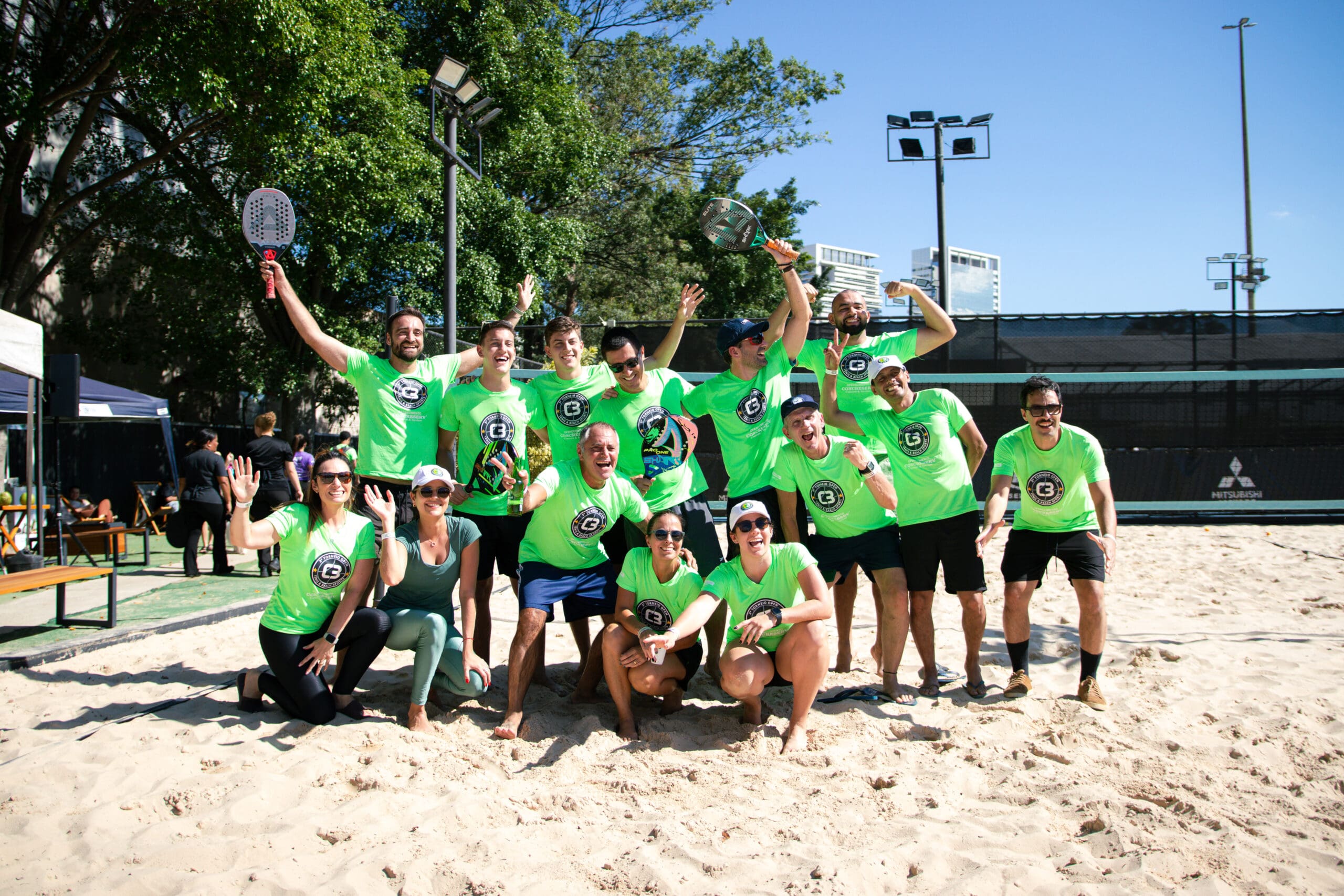 Grupo de atletas com camisetas verdes do 3º C3 Open Beach Sports posa sorridente e comemorativo na quadra de areia, celebrando o espírito esportivo do evento.