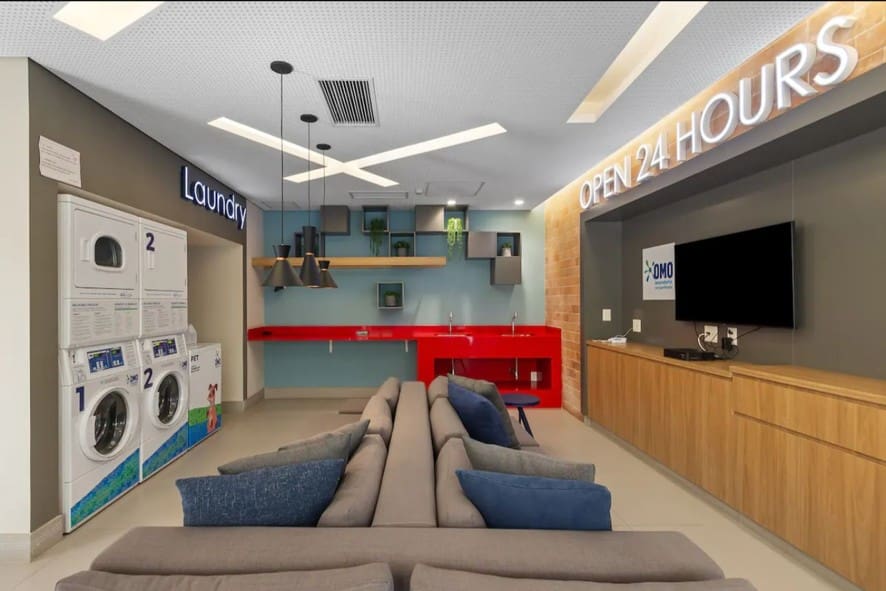 Espaço moderno de lavanderia compartilhada OMO em condomínio residencial, com máquinas de lavar e secar, área de estar com sofás, televisão e ambiente decorado, funcionando 24 horas.