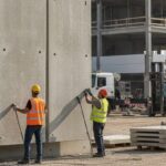 Dois operários com coletes e capacetes de segurança posicionam grandes painéis pré-moldados de concreto em um canteiro de obras, utilizando uma grua. Ao fundo, estrutura de edifício em construção e equipamentos de obra.