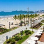 Vista aérea de uma orla revitalizada em Balneário Camboriú, Santa Catarina, mostrando calçadão moderno com quiosques, ciclovia, palmeiras, áreas de lazer e a praia ao fundo, com o mar azul e prédios altos na linha do horizonte.
