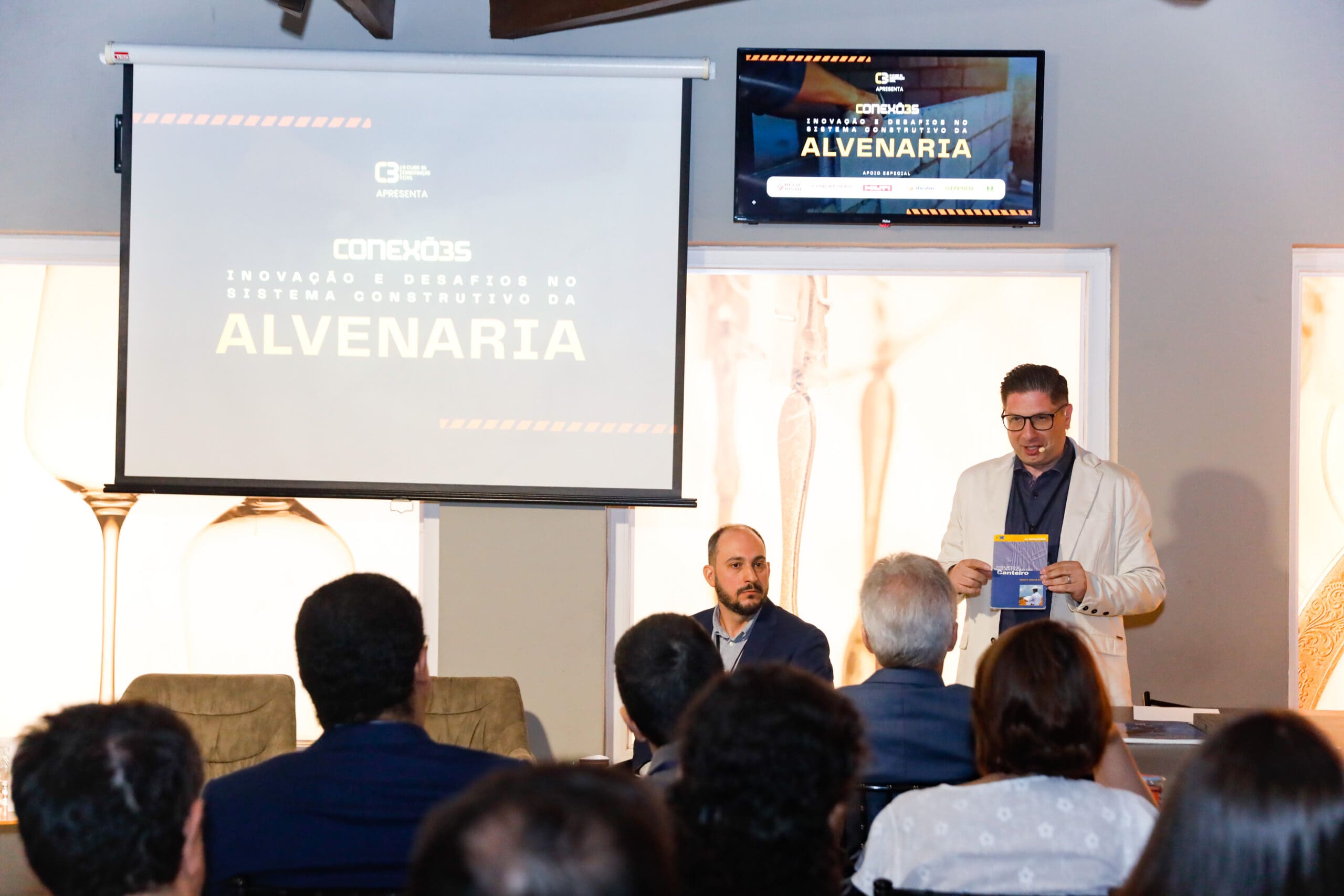Alexandre Britez mediando painel sobre inovação e desafios no sistema construtivo da alvenaria durante o evento Conexões C3, diante de uma plateia atenta.
