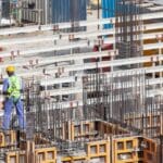 Trabalhador da construção civil com equipamentos de segurança inspeciona estrutura de concreto e aço em obra.