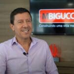 Milton Bigucci Junior sorri em uma entrevista, vestindo camisa social clara. Ao fundo, uma tela exibe o logotipo da MBigucci com a frase "Construindo uma vida melhor" e um selo comemorativo de 40 anos.