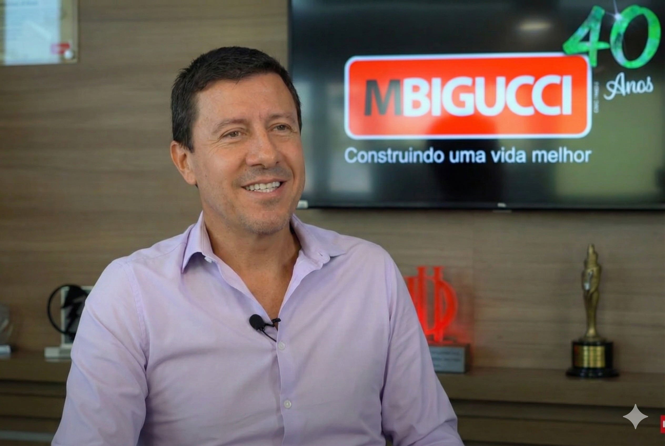 Milton Bigucci Junior sorri em uma entrevista, vestindo camisa social clara. Ao fundo, uma tela exibe o logotipo da MBigucci com a frase "Construindo uma vida melhor" e um selo comemorativo de 40 anos.