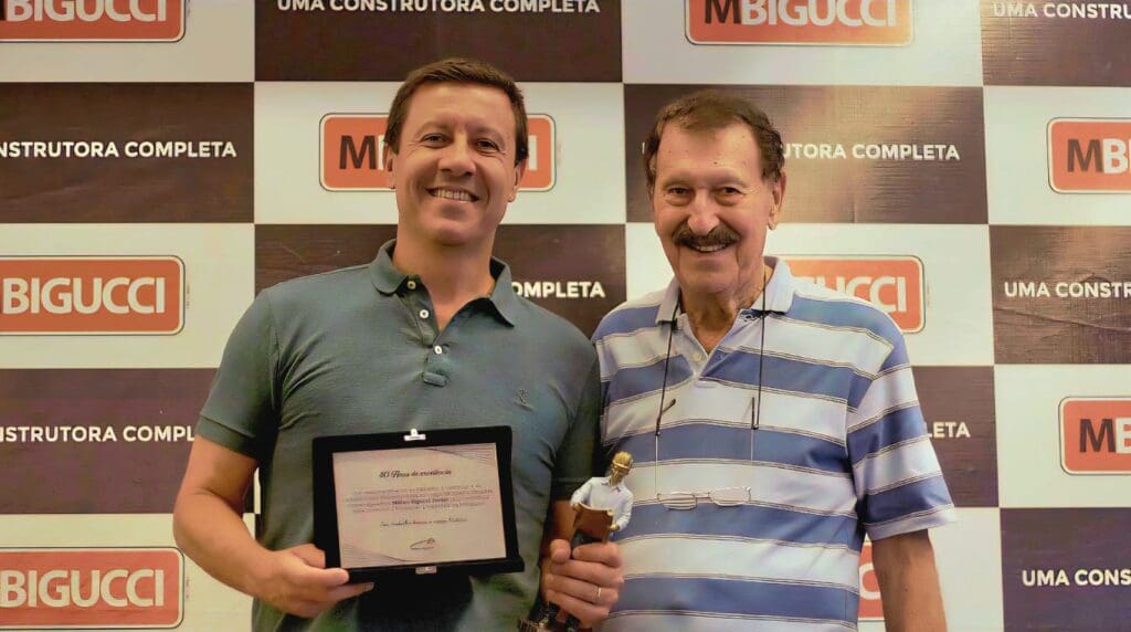 Milton Bigucci Junior, à esquerda, sorri ao lado de seu pai, Milton Bigucci, à direita. Junior segura uma placa de homenagem e um troféu em reconhecimento aos seus 40 anos de carreira na construção civil, em frente a um painel com logotipos da MBigucci.