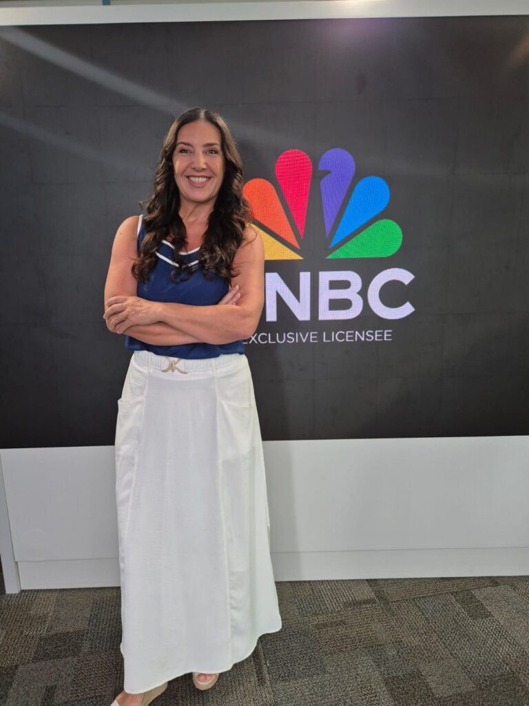 Roberta Bigucci sorrindo de braços cruzados em frente ao painel com o logotipo da NBC no estúdio da TV Times Brasil.