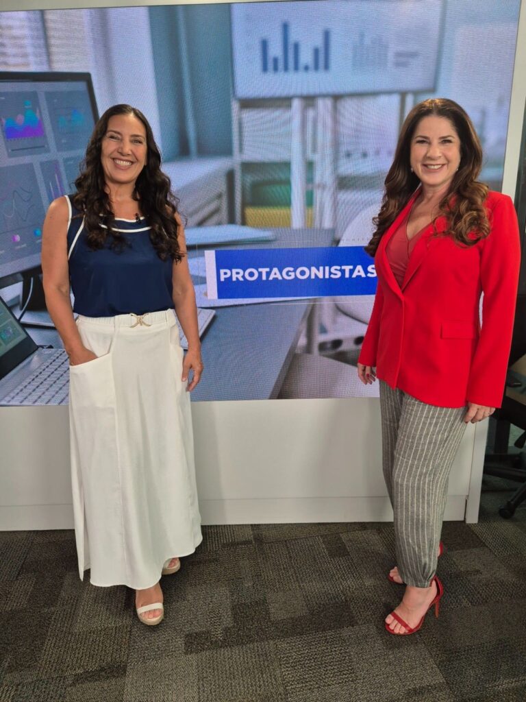 Roberta Bigucci e Christiane Pelajo sorrindo lado a lado no estúdio do programa Protagonistas, da TV Times Brasil, com painel digital ao fundo.