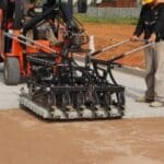 Equipamento de assentamento mecanizado instala piso paginado em espinha de peixe em obra, com operadores acompanhando o processo.