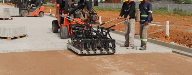 Equipamento de assentamento mecanizado instala piso paginado em espinha de peixe em obra, com operadores acompanhando o processo.