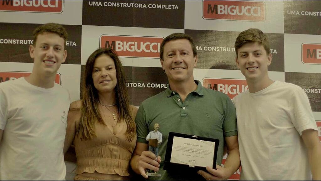 Milton Bigucci Junior sorrindo ao lado de sua esposa, Ana Ruth, e seus dois filhos, Rafael e Bruno, segurando uma placa de homenagem e um troféu pelos seus 40 anos de carreira na construção civil.