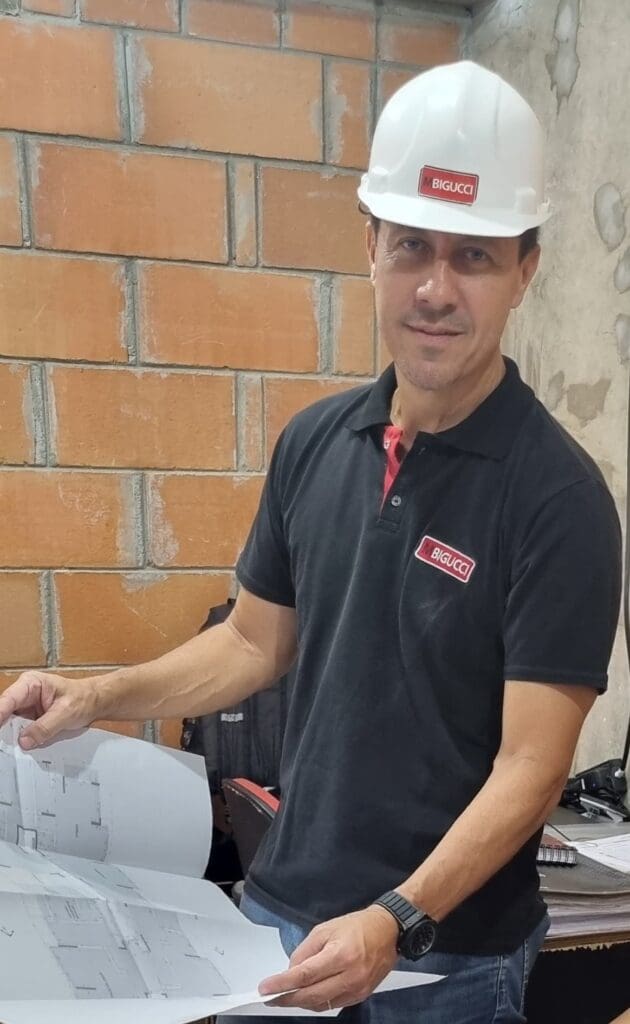 Diretor técnico Milton Bigucci Junior, utilizando capacete branco e camisa polo com o logotipo da MBigucci, analisa projetos de engenharia em um canteiro de obras.