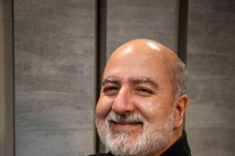 Paulo Oliveira, engenheiro civil e referência em industrialização da construção, sorrindo com os braços cruzados em ambiente corporativo.