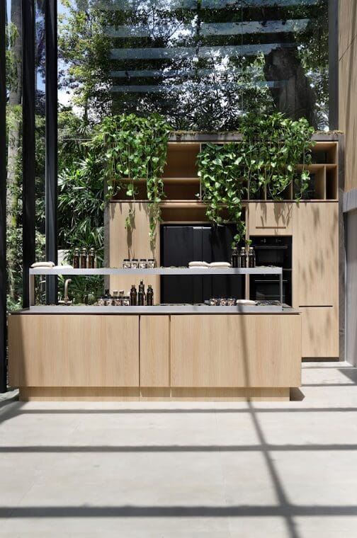 Cozinha contemporânea da SCA Jardim Europa com armários em madeira clara, ilha central e integração com área verde externa, destacando design sofisticado e elementos naturais.