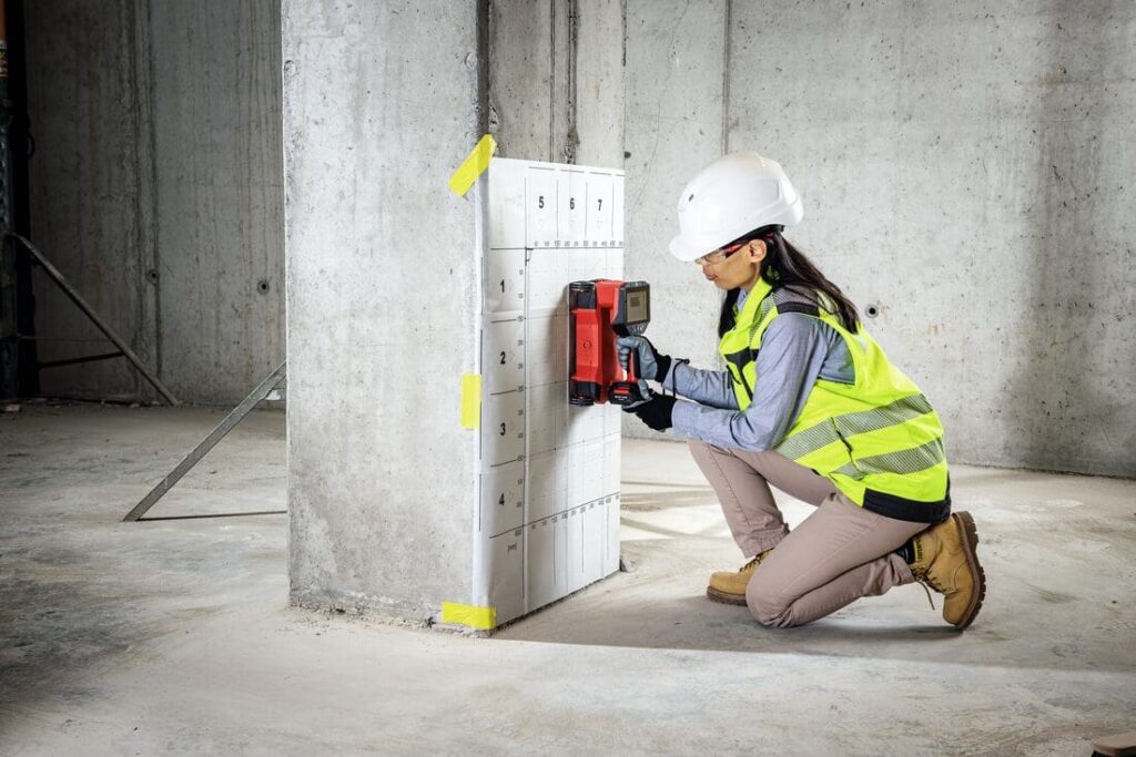 Engenheira utiliza scanner Hilti para inspeção estrutural em pilar de concreto durante obra de retrofit, vestindo EPI completo.