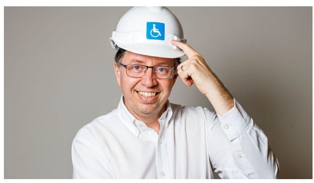 Eduardo Ronchetti de Castro sorrindo e apontando para um capacete branco de obra com o símbolo internacional de acessibilidade, em fundo neutro cinza.