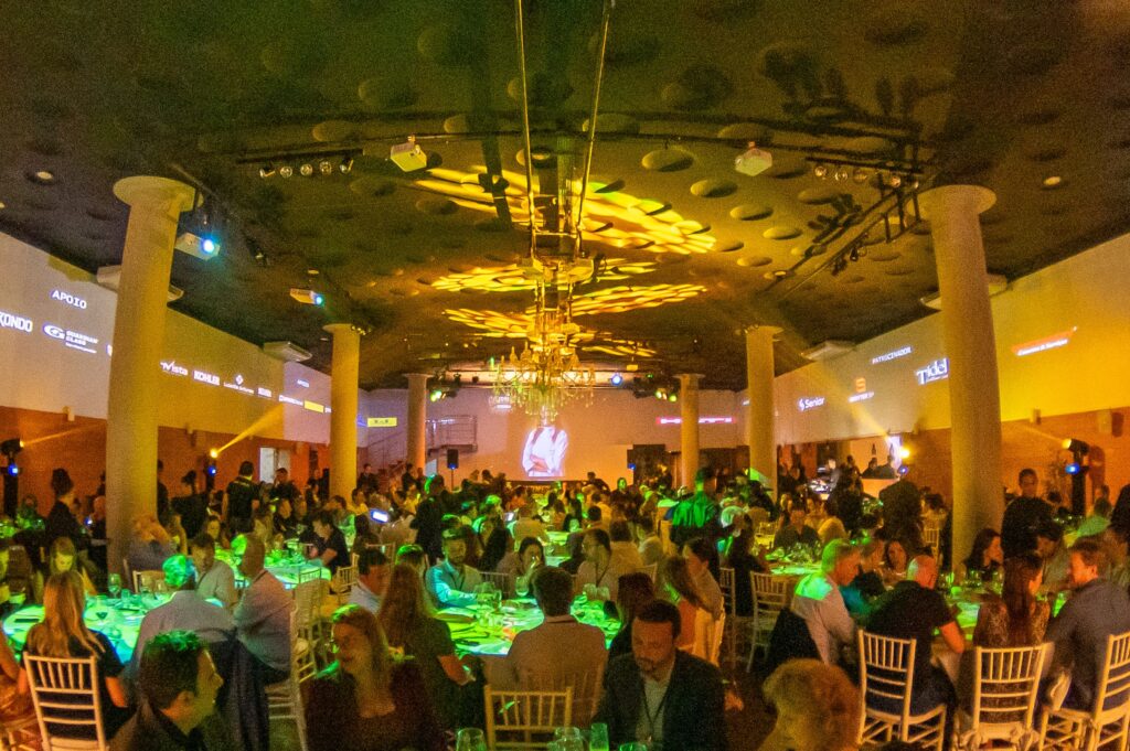 Vista panorâmica do salão do Gourmet Immersive Experience do C3 – O Clube da Construção Civil completamente lotado, com centenas de convidados em mesas redondas, iluminação cênica em verde e âmbar, lustre de cristal central, telão com imagem da chef ao fundo e logos de patrocinadores como Kohler, InVista, Guardian Glass e Senior nas paredes