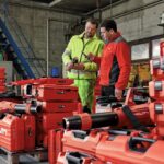 Dois profissionais analisam ferramentas Hilti em um canteiro de obras, cercados por equipamentos e maletas vermelhas da marca, destacando tecnologia e organização no setor da construção civil.