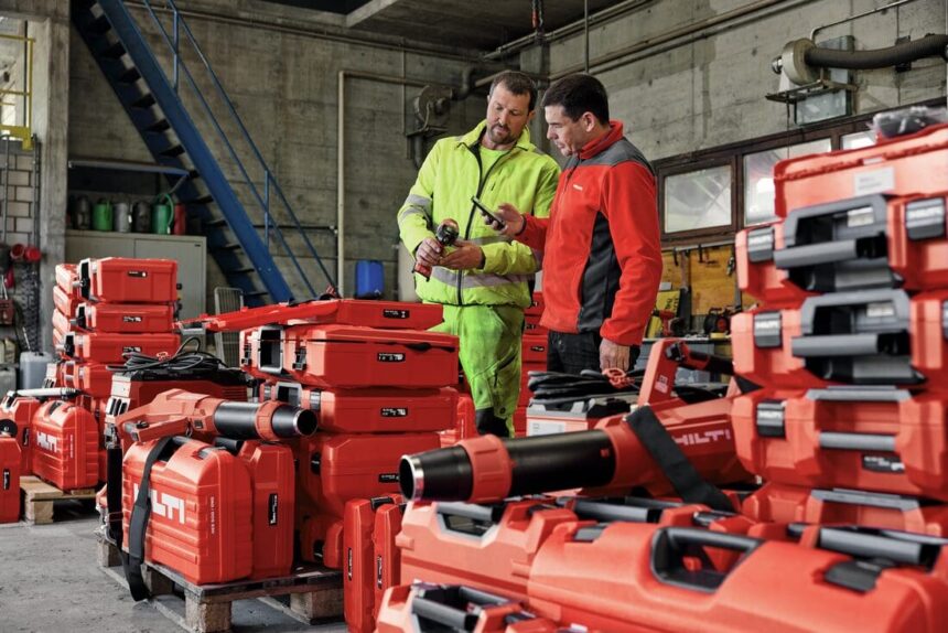 Dois profissionais analisam ferramentas Hilti em um canteiro de obras, cercados por equipamentos e maletas vermelhas da marca, destacando tecnologia e organização no setor da construção civil.