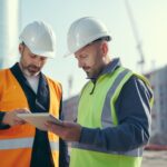Dois engenheiros de obras civis usando capacete e colete de segurança analisam dados em um tablet no canteiro de obras, com edifícios em construção ao fundo.