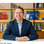 Eduardo Ronchetti de Castro, arquiteto e consultor referência nacional em acessibilidade arquitetônica, sentado à mesa de trabalho com plantas técnicas, capacete com símbolo de acessibilidade e livros sobre o tema ao fundo.