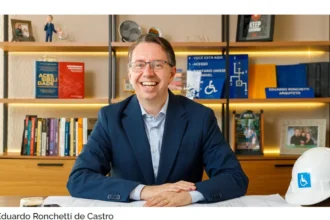 Eduardo Ronchetti de Castro, arquiteto e consultor referência nacional em acessibilidade arquitetônica, sentado à mesa de trabalho com plantas técnicas, capacete com símbolo de acessibilidade e livros sobre o tema ao fundo.
