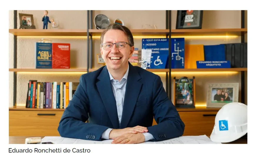 Eduardo Ronchetti de Castro, arquiteto e consultor referência nacional em acessibilidade arquitetônica, sentado à mesa de trabalho com plantas técnicas, capacete com símbolo de acessibilidade e livros sobre o tema ao fundo.