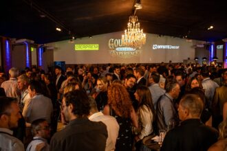 Convidados do setor da construção civil participam do coquetel de recepção do Gourmet Immersive Experience C3 no Villa Glam, com ambiente sofisticado, iluminação especial e logomarcas de patrocinadores projetadas nas paredes.