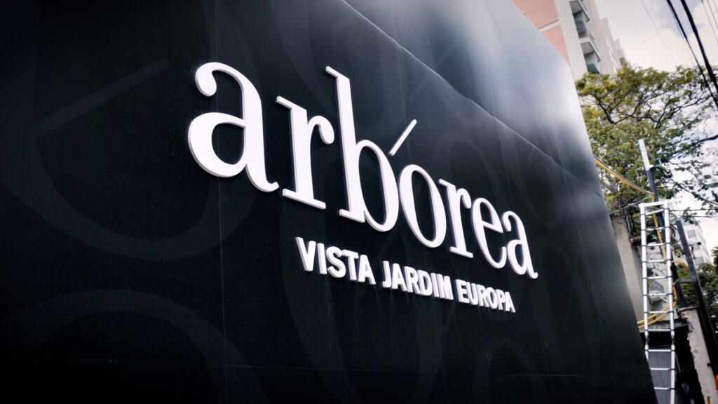 Placa de identificação do empreendimento Arbórea Vista Jardim Europa em destaque na fachada do canteiro de obras, com letras brancas em fundo preto e edifícios ao fundo.