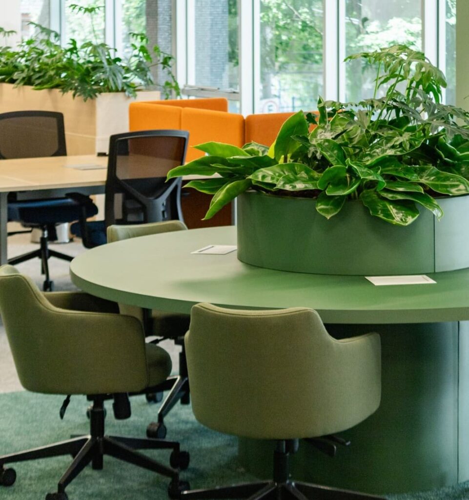 Espaço de escritório colaborativo com mesa redonda verde, cadeiras giratórias, plantas naturais no centro e ao fundo, além de sofás laranja e grandes janelas com vista para áreas verdes externas.