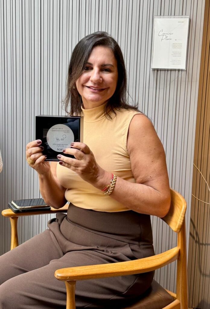 Márcia Dias, da SCA Jardim Europa, sentada e sorrindo, segura o troféu de Melhor Gerente recebido na premiação TOPS 2025.