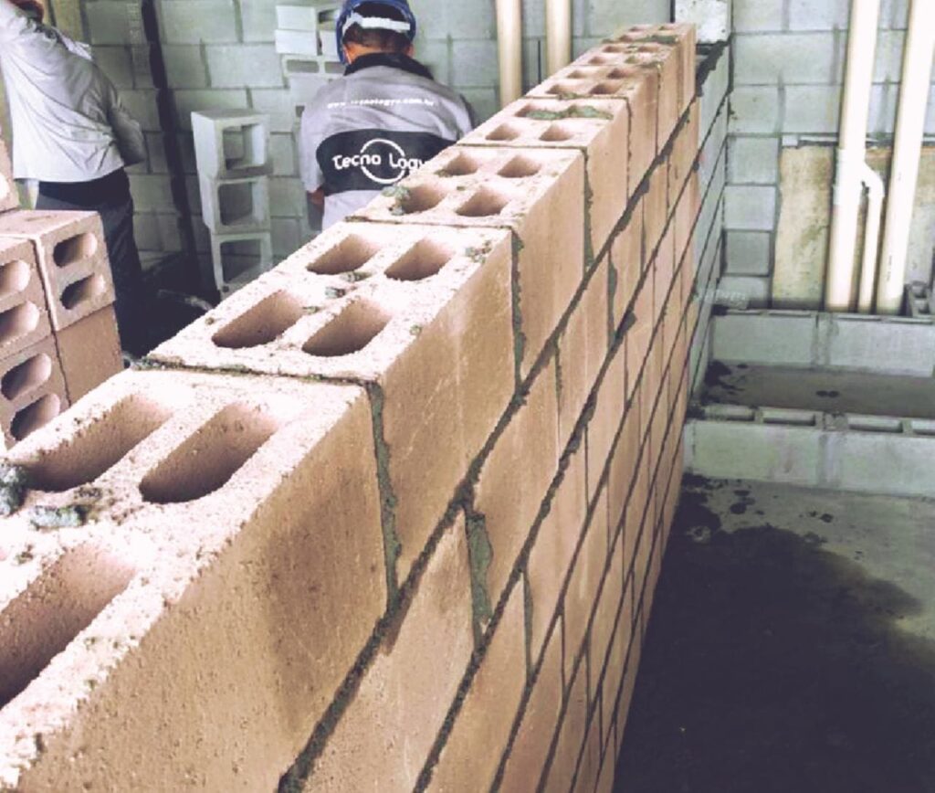 Detalhe de parede em construção com blocos cerâmicos estruturais, com profissionais trabalhando ao fundo e instalações hidráulicas aparentes.