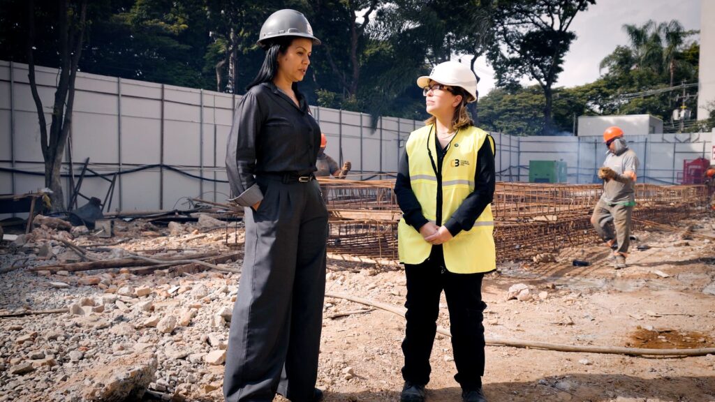 Bárbara Martinez, do C3, entrevista Bianca Lopes, diretora da Servter SP, usando capacete e equipamentos de segurança, conversam no canteiro de obras do Arbórea Vista Jardim Europa, com estruturas de aço e trabalhadores ao fundo.