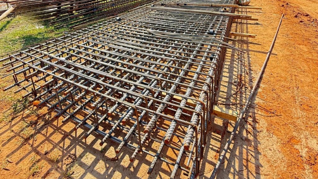 Estrutura de armação de aço pronta para fundação, posicionada sobre solo de terra em canteiro de obras, evidenciando organização e precisão no preparo estrutural.