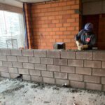 Operário assentando blocos estruturais de concreto em obra residencial, com paredes de tijolos ao fundo, demonstrando técnicas modernas de alvenaria.