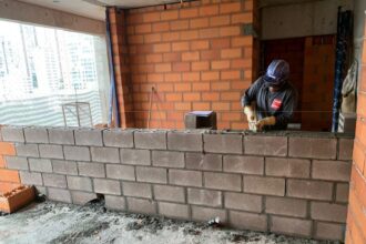 Operário assentando blocos estruturais de concreto em obra residencial, com paredes de tijolos ao fundo, demonstrando técnicas modernas de alvenaria.